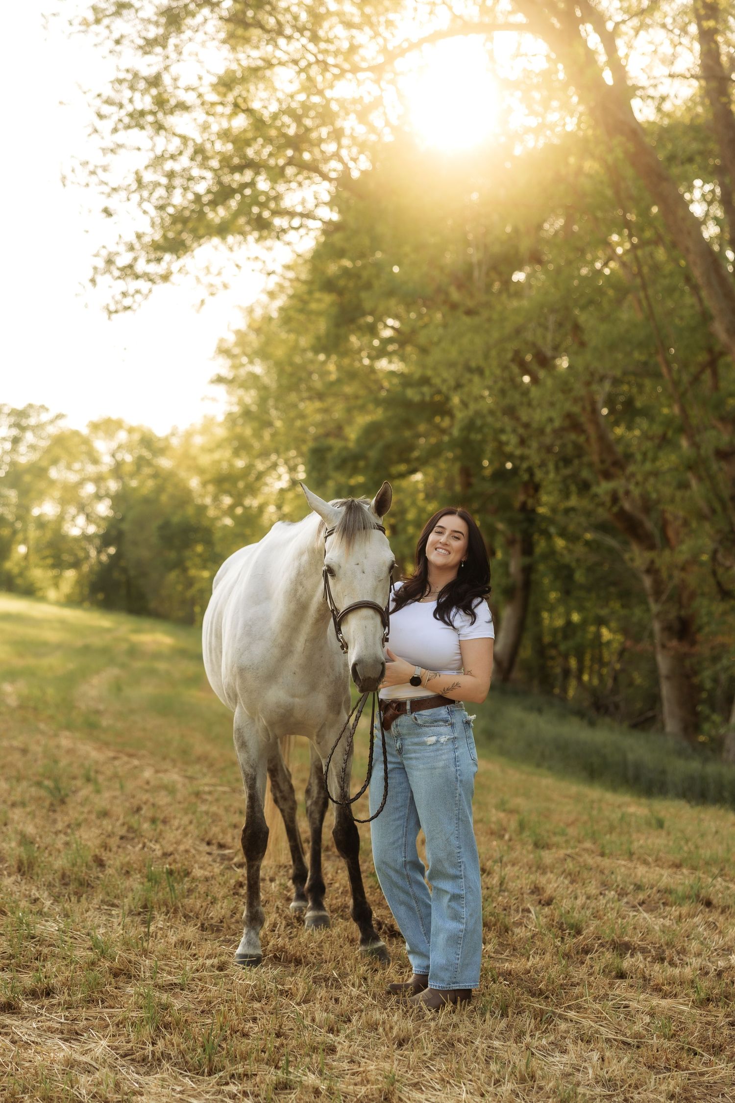 Smiling woman in white top and jeans poses with a white horse in a sunny pastoral field at sunset.