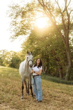 Smiling brunette woman in jeans standing beside a gray horse in a sunlit green meadow at sunset.