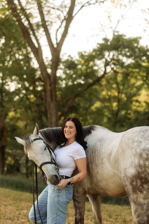Smiling brunette woman in white t-shirt and jeans posing with a gray horse in golden sunset light.