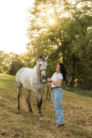 Woman in white top and jeans leading a gray horse through a sunlit meadow with trees in background.
