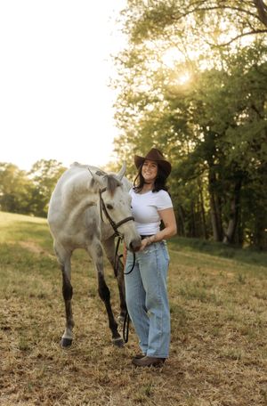 Young woman in cowboy hat and jeans smiles while leading a white horse in a sunlit green pasture.