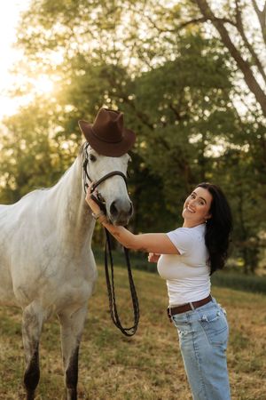Smiling woman holding a brown cowboy hat on a gray horse in a sun-drenched outdoor setting.