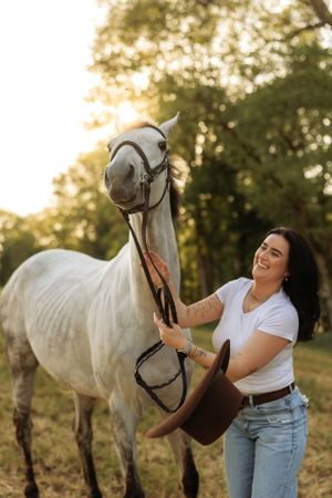 Laughing woman placing a brown cowboy hat on a gray horse during a warm golden sunset outdoors.