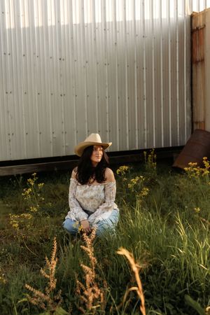 Young woman in cowboy hat and floral off-shoulder top sits in green field beside rustic metal barn wall.