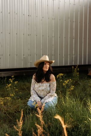 Brunette woman in cowboy hat and floral top sitting in tall grass near a white barn at golden hour.