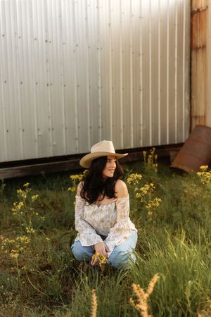 Brunette woman in floral off-shoulder top and cowboy hat sits in wildflowers beside a rustic barn.