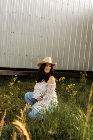 Brunette woman wearing cowboy hat and floral top sits among wildflowers in front of corrugated metal wall.