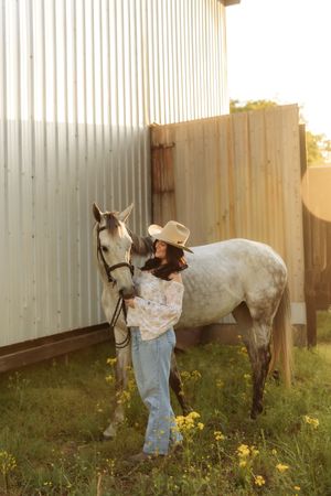 Woman in floral off-shoulder top and cowboy hat gently embracing a dapple gray horse by a barn.