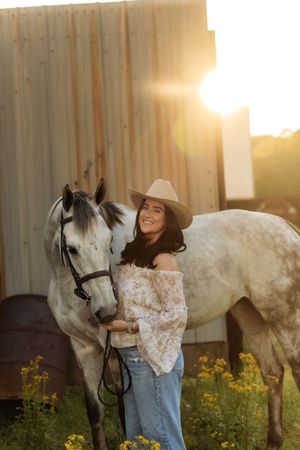 Smiling woman in floral blouse and tan cowboy hat standing beside a gray horse near a barn at sunset.