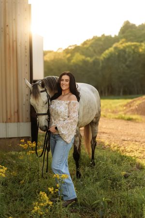 Woman in floral top and jeans smiles beside a gray horse near a wooden barn at golden hour.