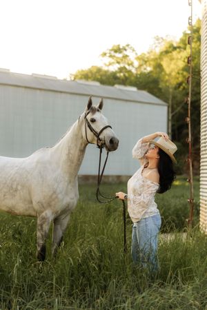 Young woman in lace top and cowboy hat poses beside a white horse near a barn in golden sunlight.