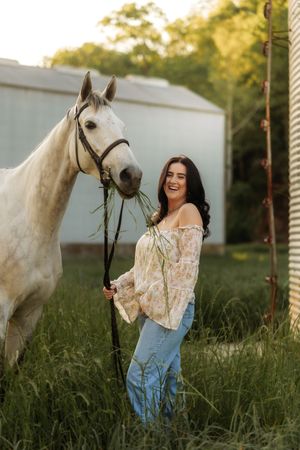Laughing woman in floral blouse holding reins as gray horse eats grass near a rustic farm silo.