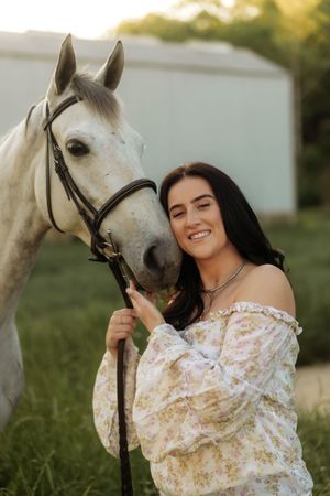 Smiling brunette woman in floral off-shoulder top posing cheek-to-cheek with a white horse at golden hour.