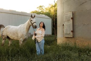 Young woman leads a white horse through green grass beside a metal grain silo at sunset.