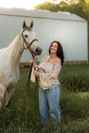 A smiling young woman in a floral top and jeans holds the reins of a white horse in a green field.