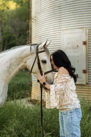 Woman in floral off-shoulder top lovingly nuzzling a dapple gray horse beside a metal grain silo.