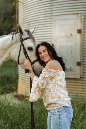 Happy young woman in floral blouse embracing a white horse near a rustic grain silo in a lush green field.