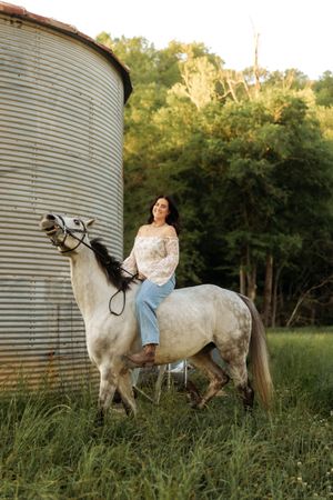 Woman in white floral top and jeans rides gray horse beside grain silo at golden hour.