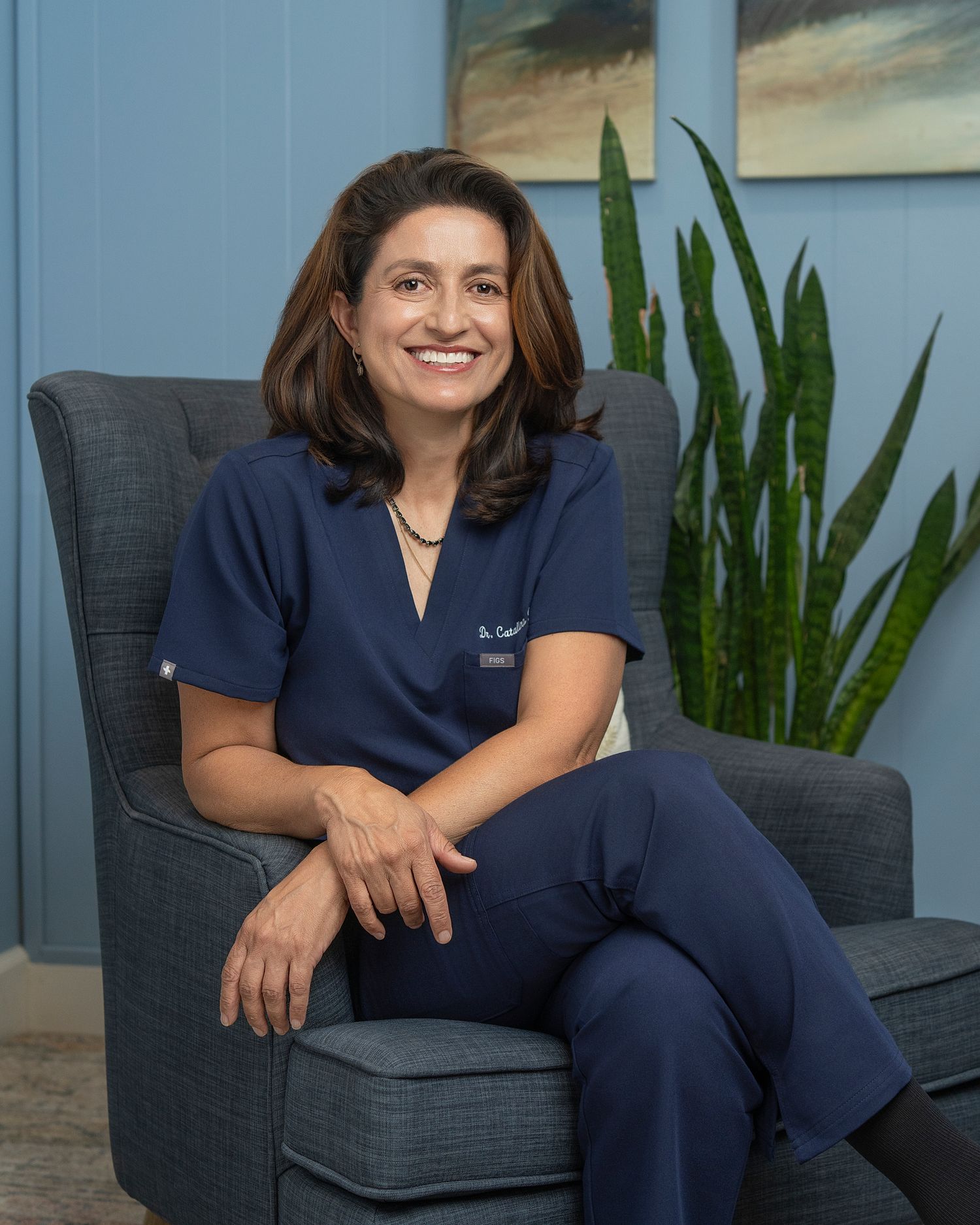 A smiling female doctor in navy scrubs sits in a gray chair beside a snake plant in a modern medical office.