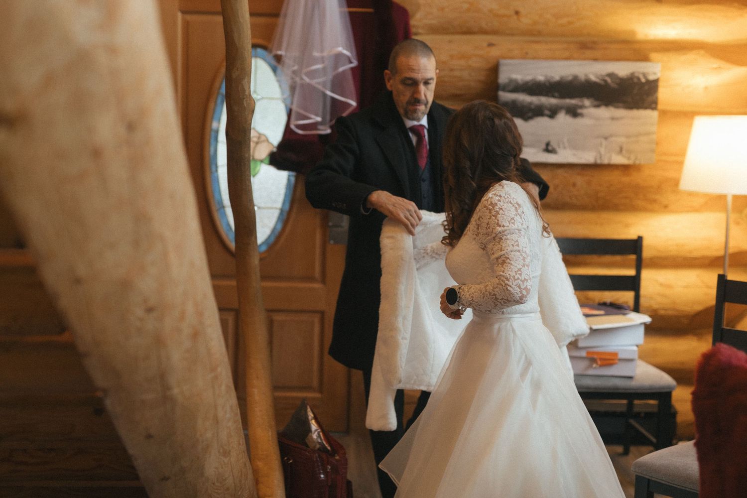 groom helps bride put on her fur coat in a cedar log cabin in Revelstoke, BC as they prepare to leave for their elopement