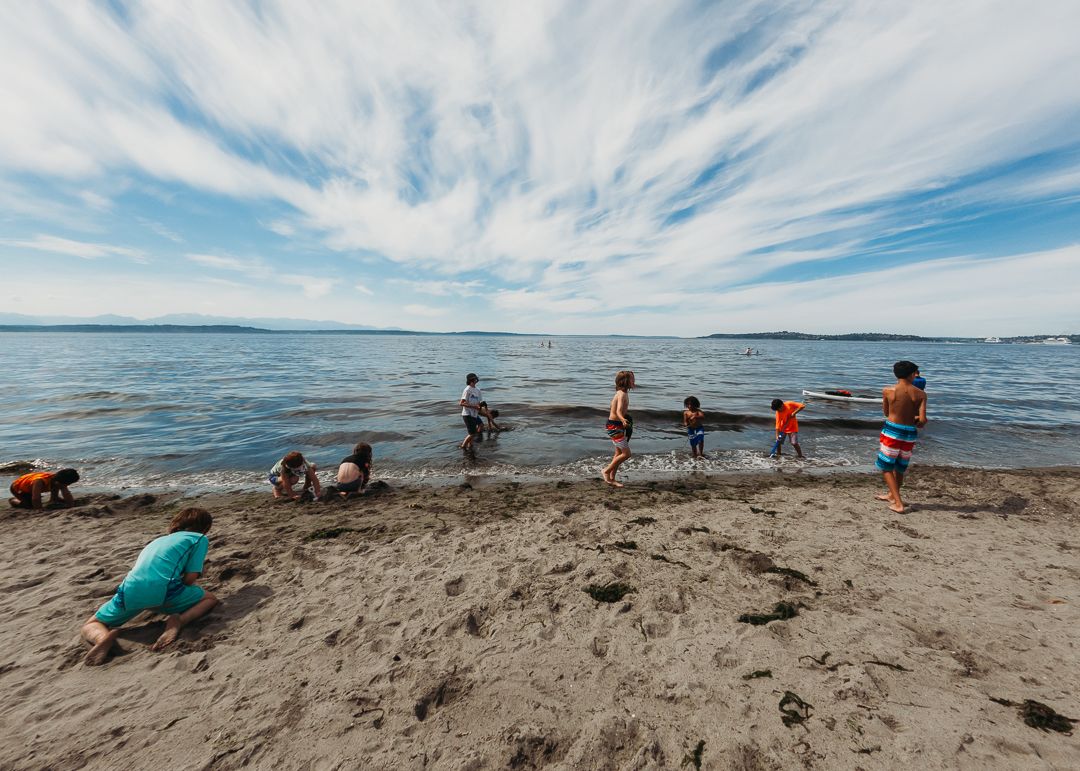 Children playing on a sandy beach, some wading in calm ocean water under a dramatic cloudy blue sky.
