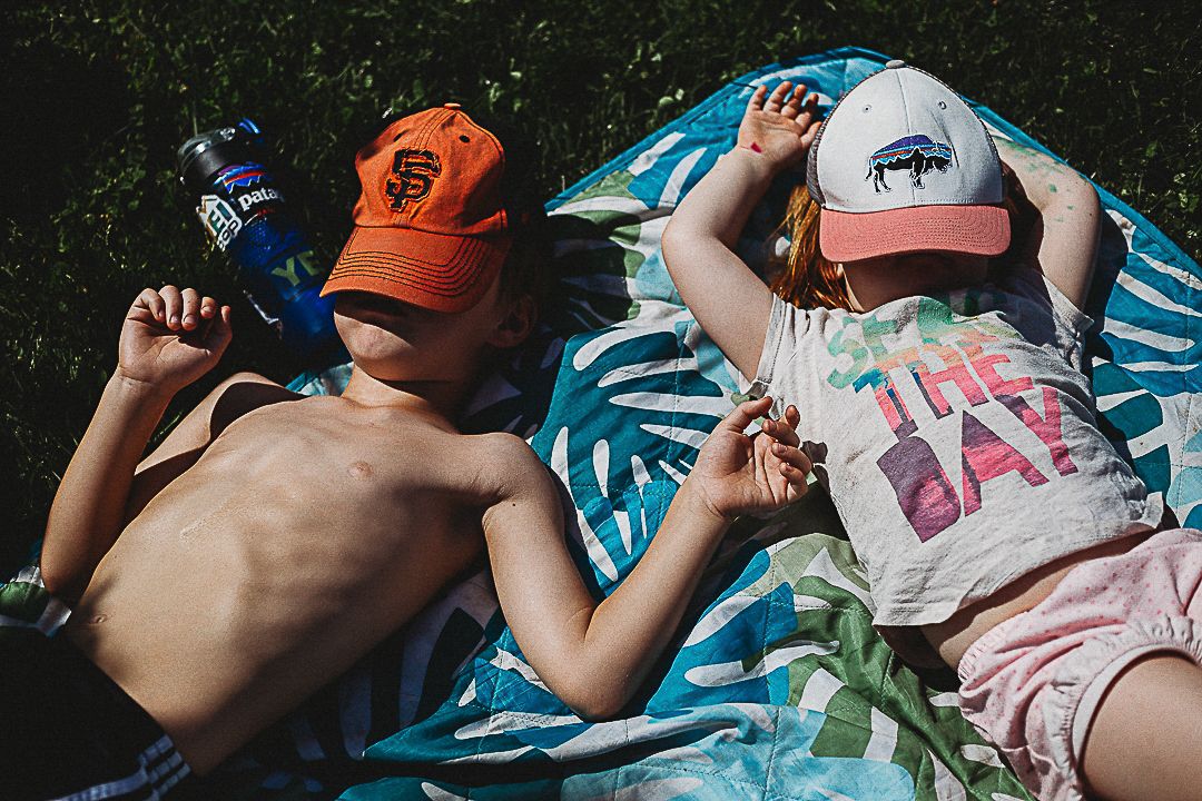 Two children lying on a tropical print blanket outdoors, wearing baseball caps and enjoying a sunny summer day.