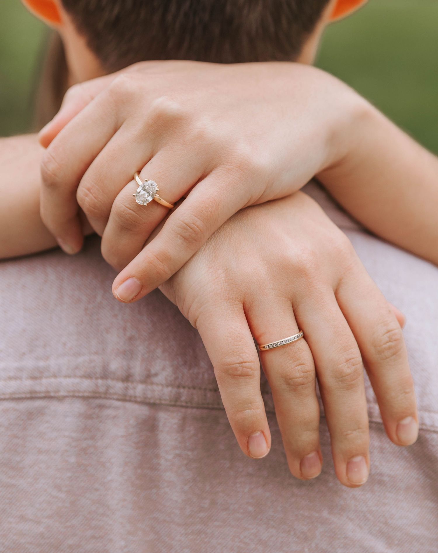 Close-up of a woman's hands resting on a man's shoulder, wearing a diamond engagement ring and a gold wedding band.