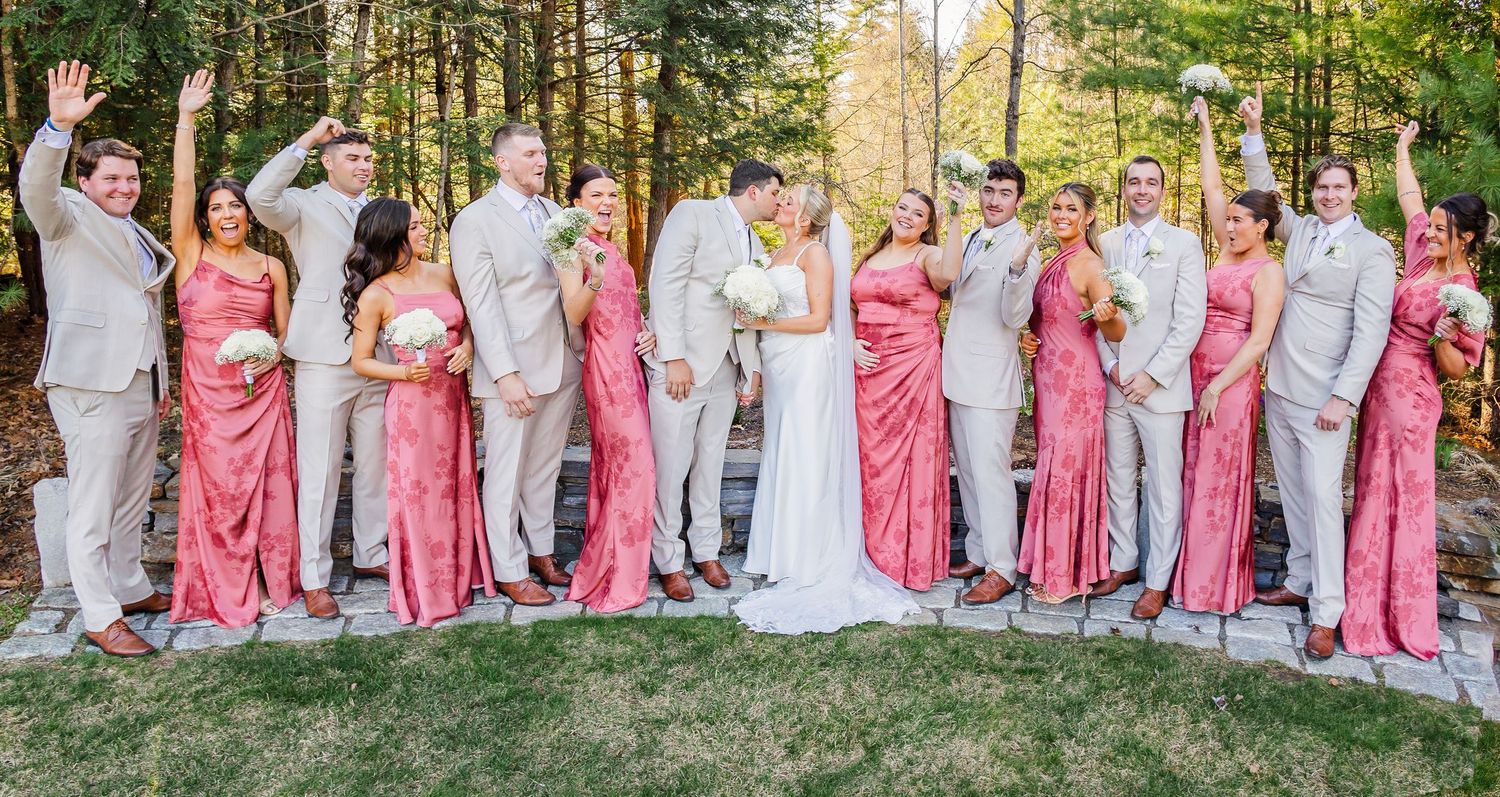 Wedding party in coral bridesmaid dresses and tan suits celebrating outdoors among trees, bride in white gown centered.