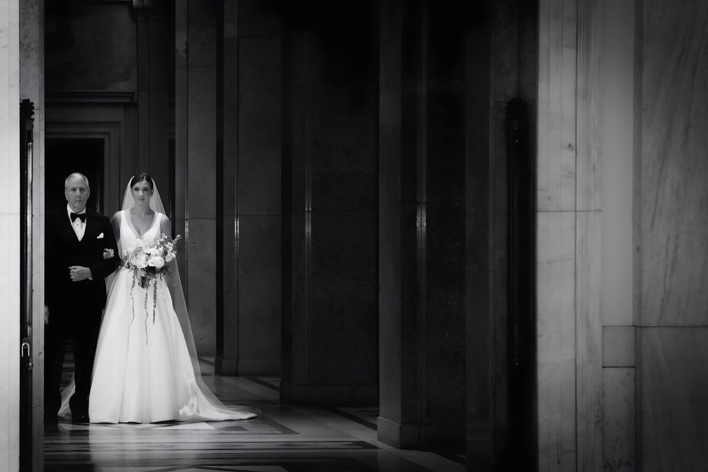 Black and white photo of bride in white gown and escort in tuxedo standing in elegant columned hallway.