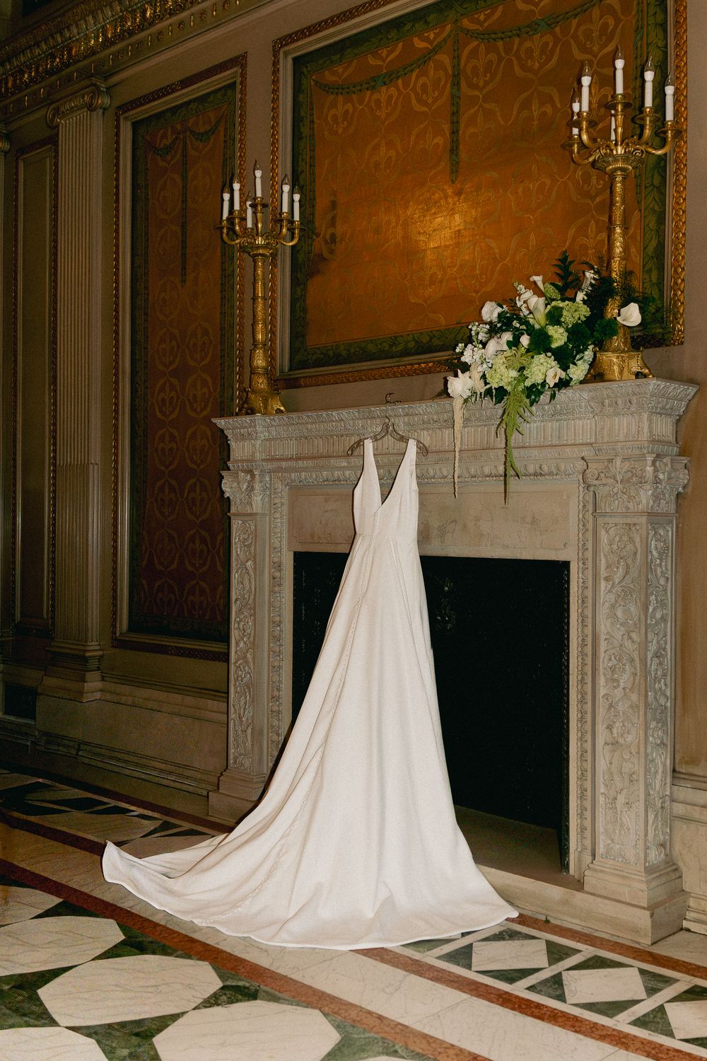 Elegant white wedding gown with long train displayed on ornate marble fireplace in luxurious gold-adorned ballroom.