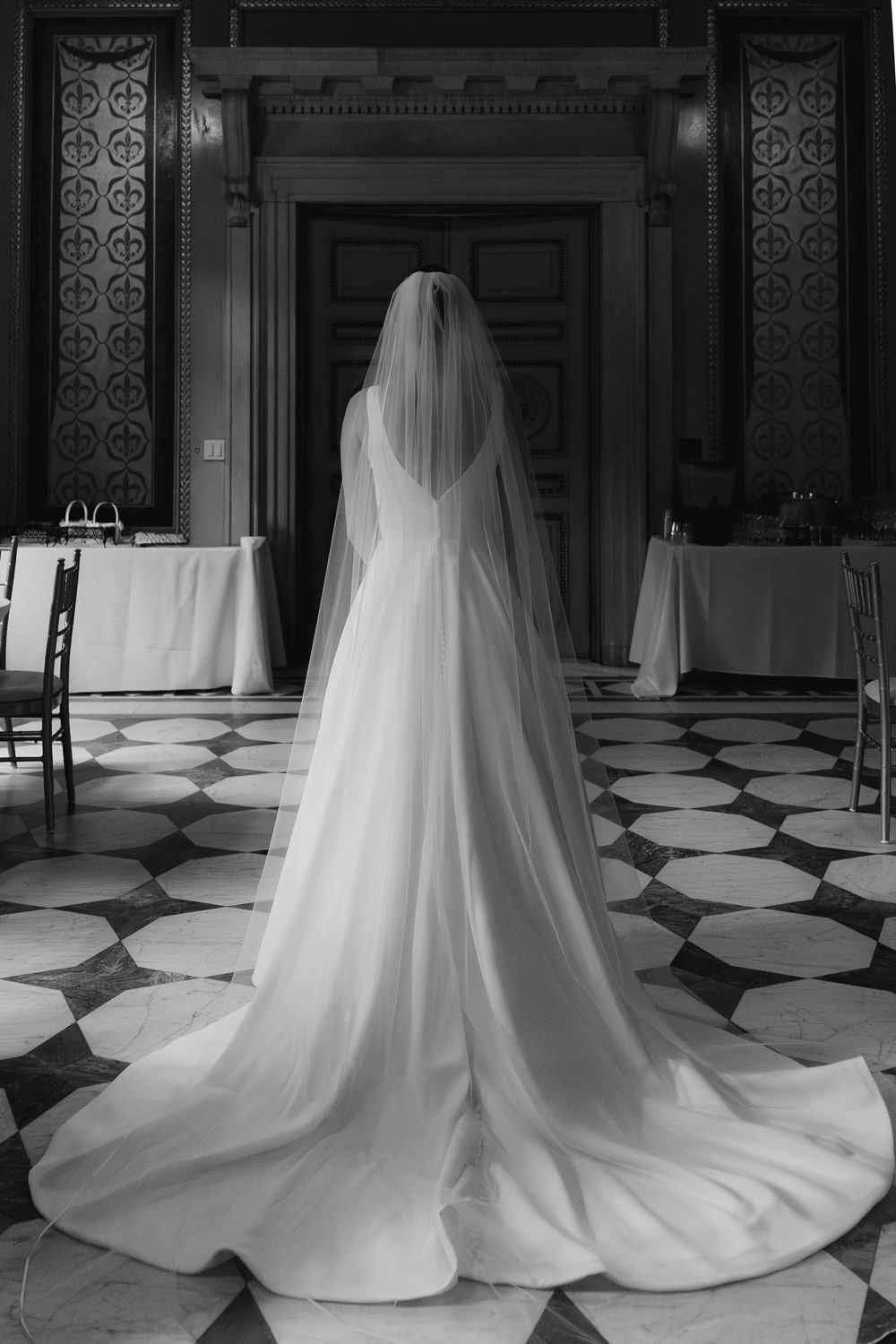 Black and white photo of a bride in a flowing white gown and veil standing in an elegant marble-floored hall.