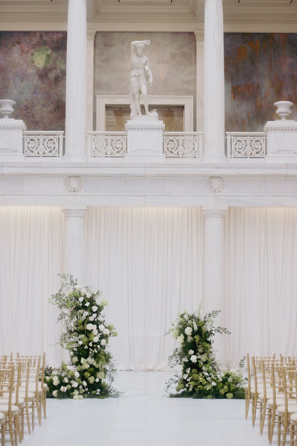 Elegant wedding ceremony setup with white floral arrangements, draped backdrop, gold chairs, and a classical statue above.