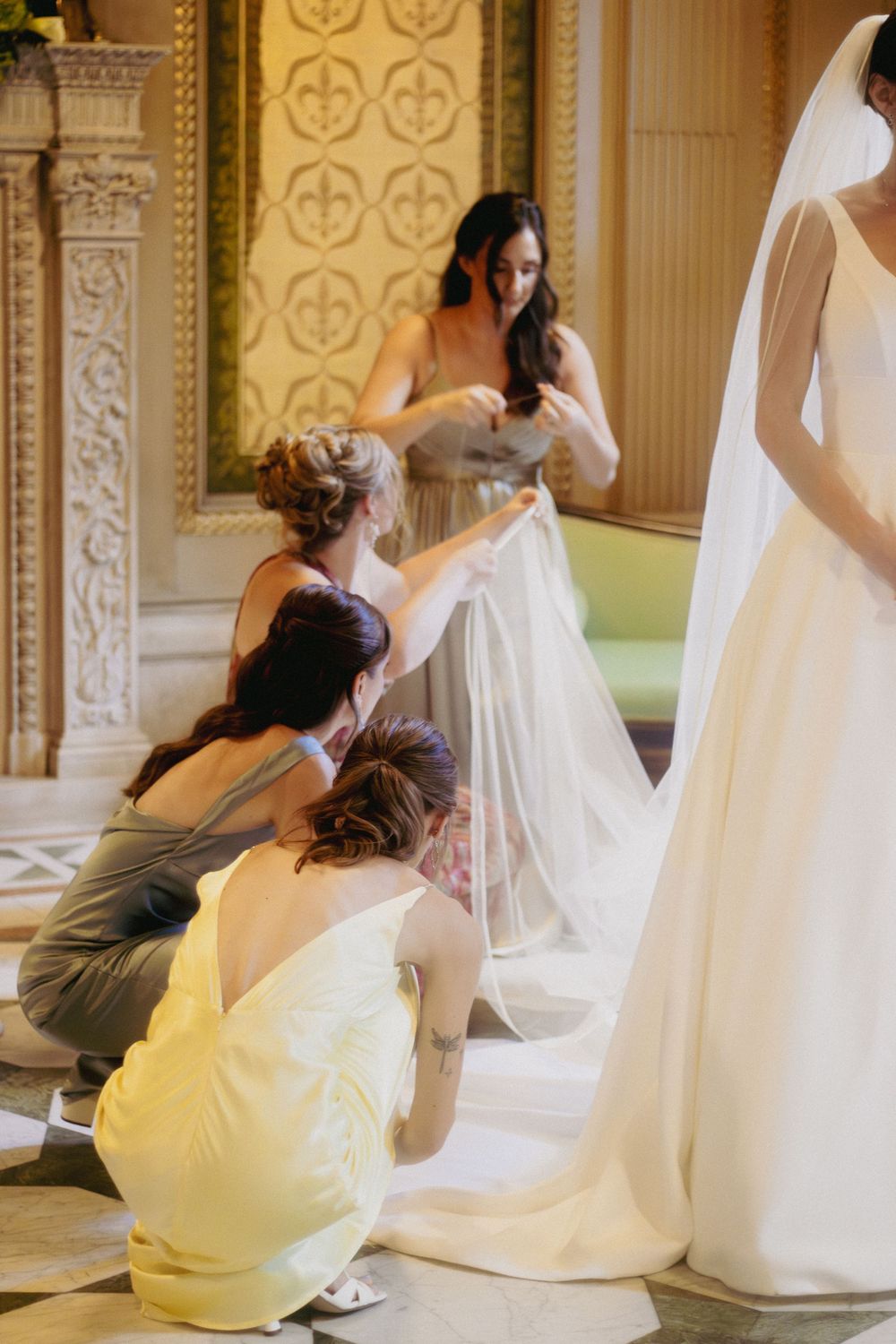 Bridesmaids kneel to arrange the train of a white wedding gown in an ornate golden venue.
