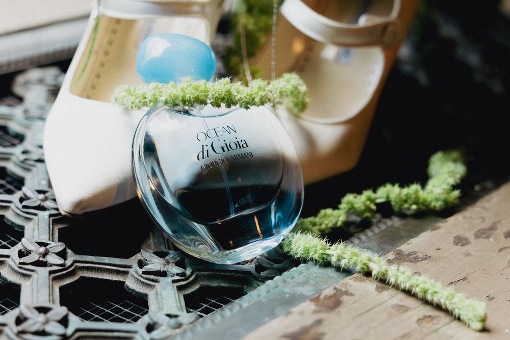 Ocean Di Gioia perfume bottle with blue liquid rests on decorative surface with green moss and white heels.