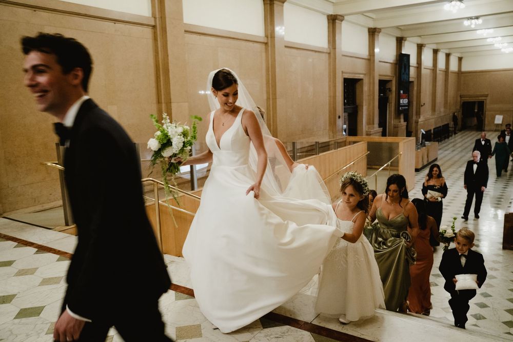 Bride in elegant white gown and veil descends grand staircase with bridesmaids at wedding venue.