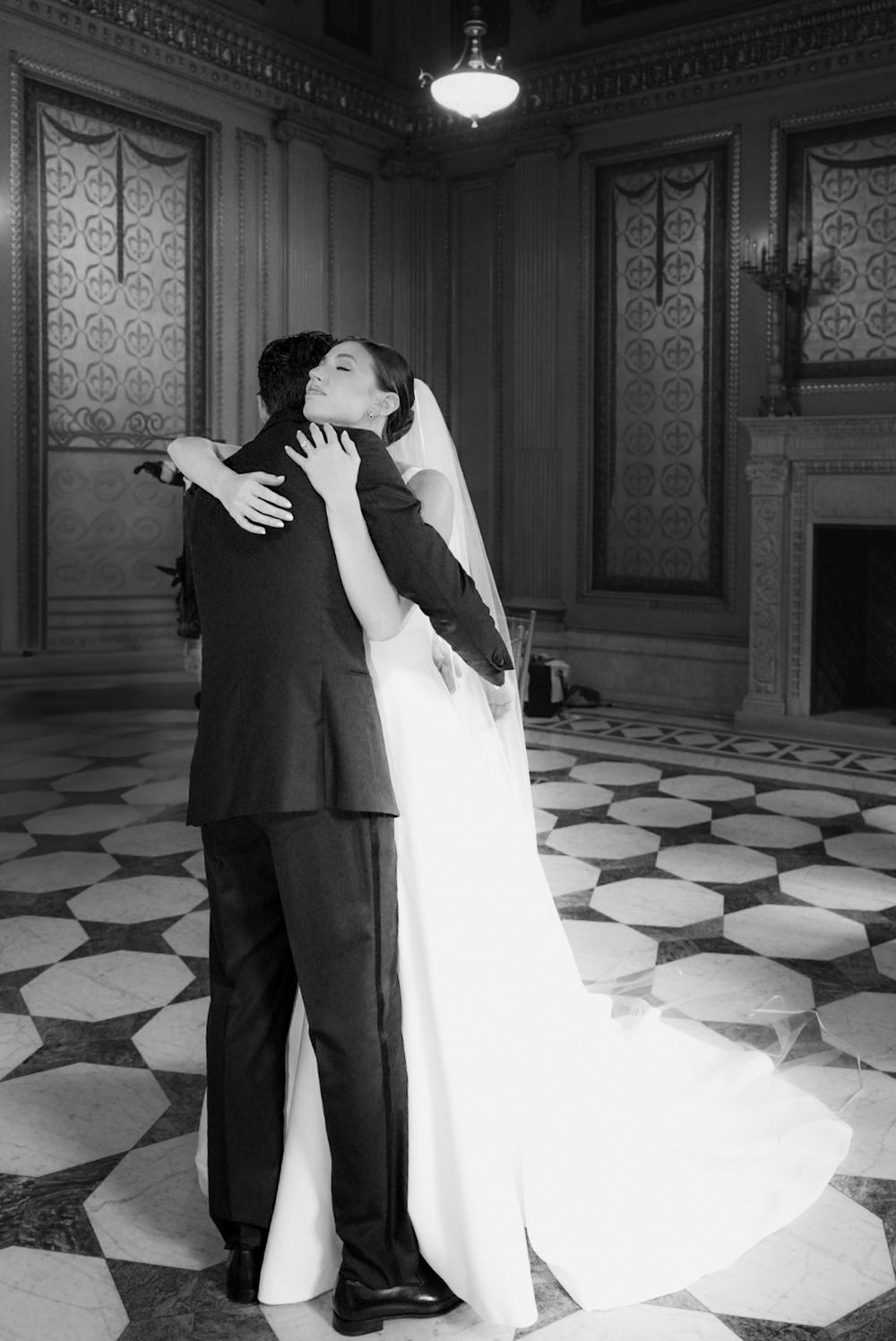 Black and white photo of a bride and groom sharing a romantic embrace in an elegant marble-floored hall.