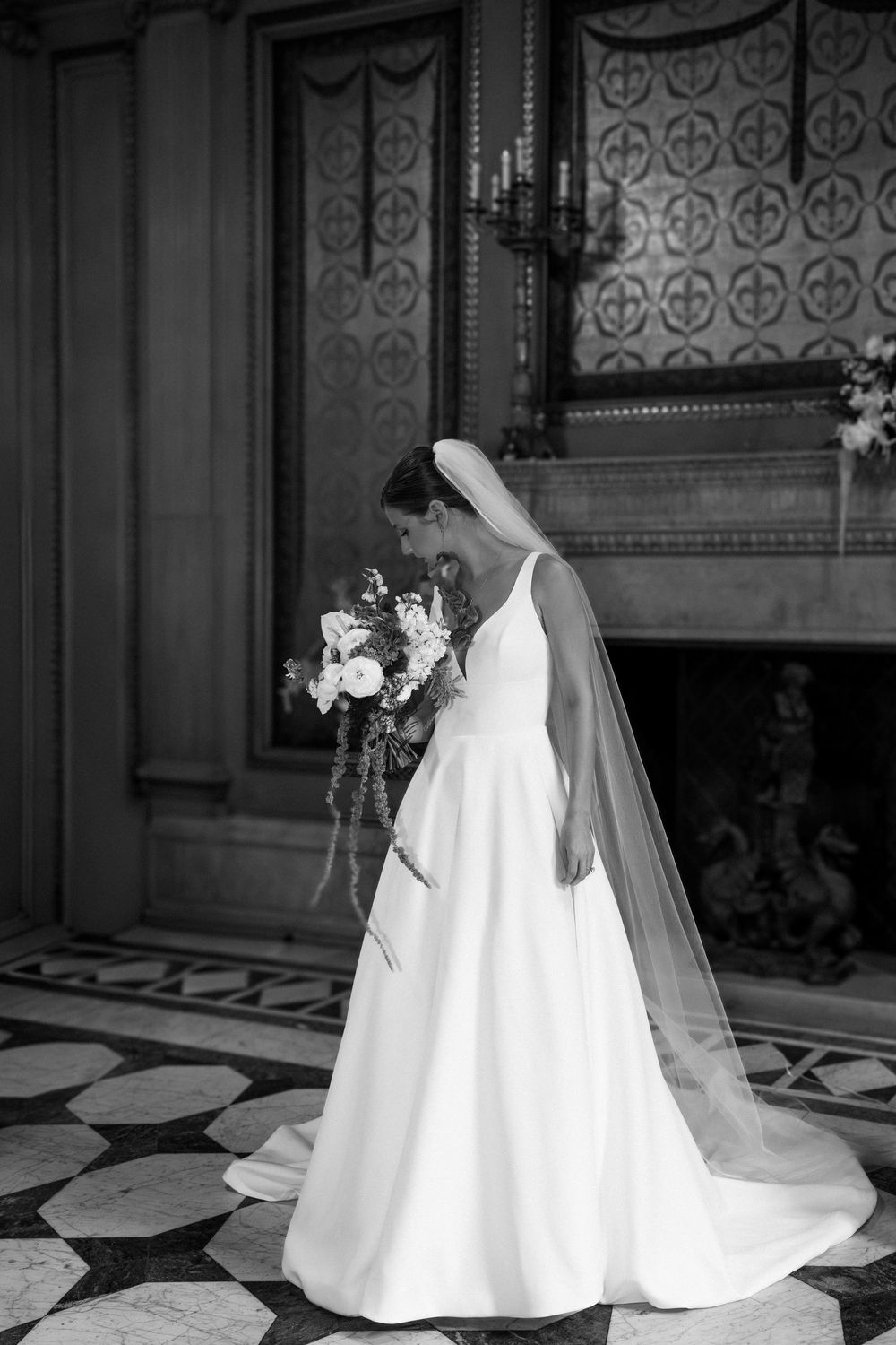 Bride in white gown and veil holds floral bouquet in ornate black and white marble hall.
