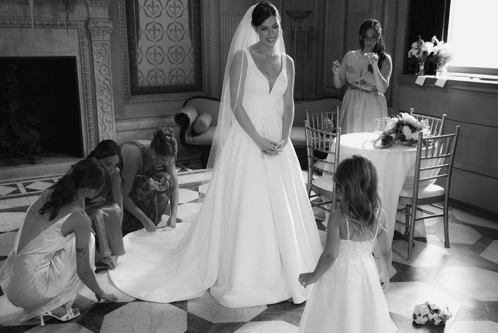 Black and white photo of a bride in a white gown and veil surrounded by bridesmaids and a flower girl in an elegant venue.
