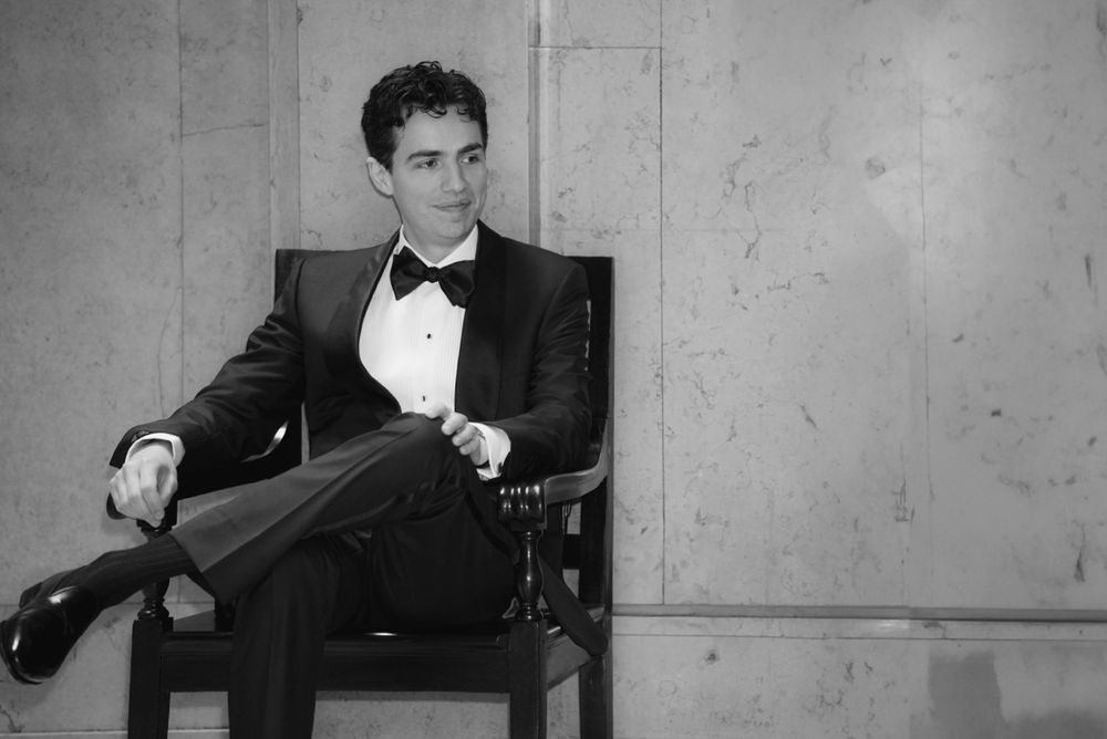 Elegant man in black tuxedo and bow tie sitting relaxed in a chair, black and white formal portrait photo.