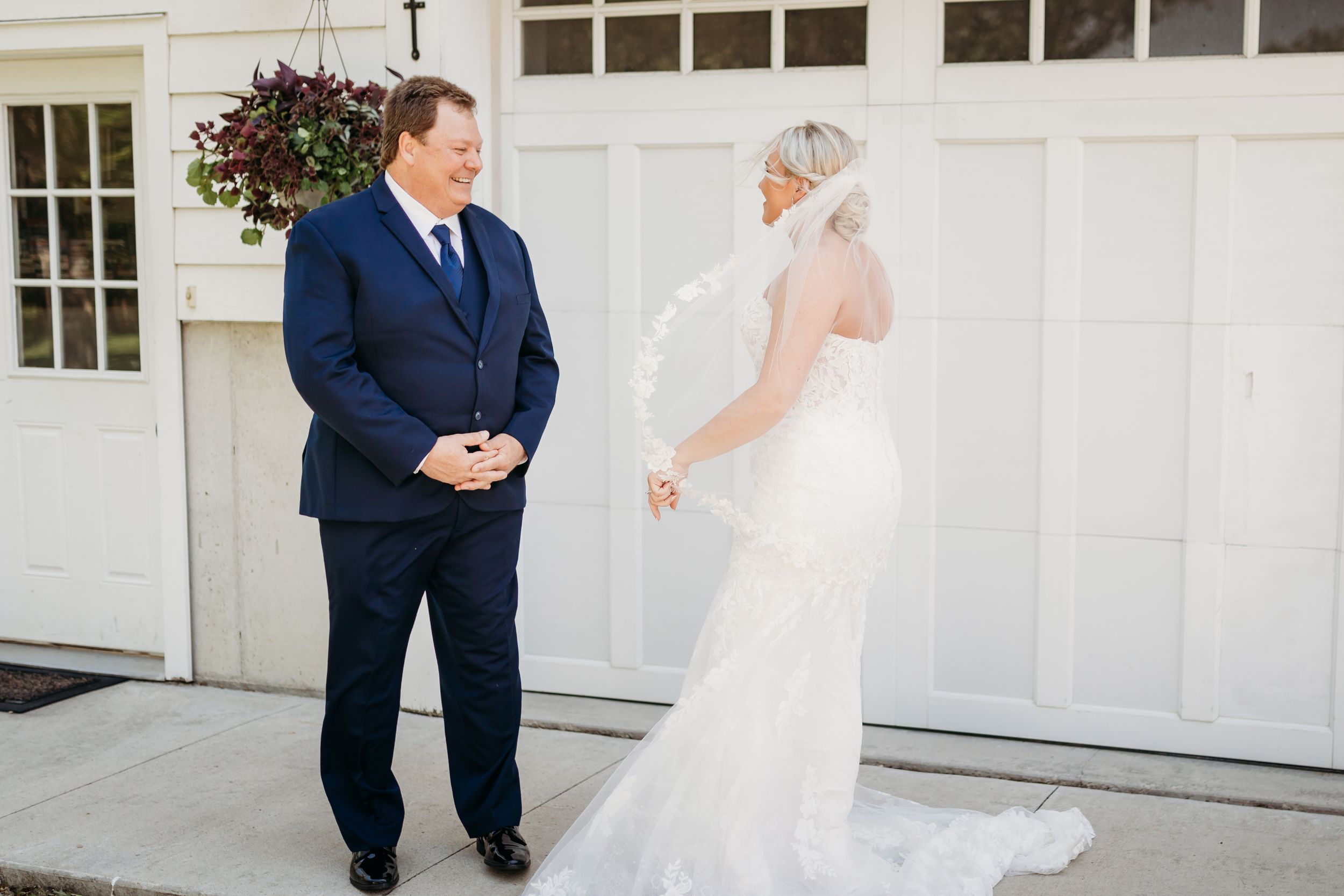 A groom in a navy suit smiles at his bride in a white lace gown and veil during a first look moment.