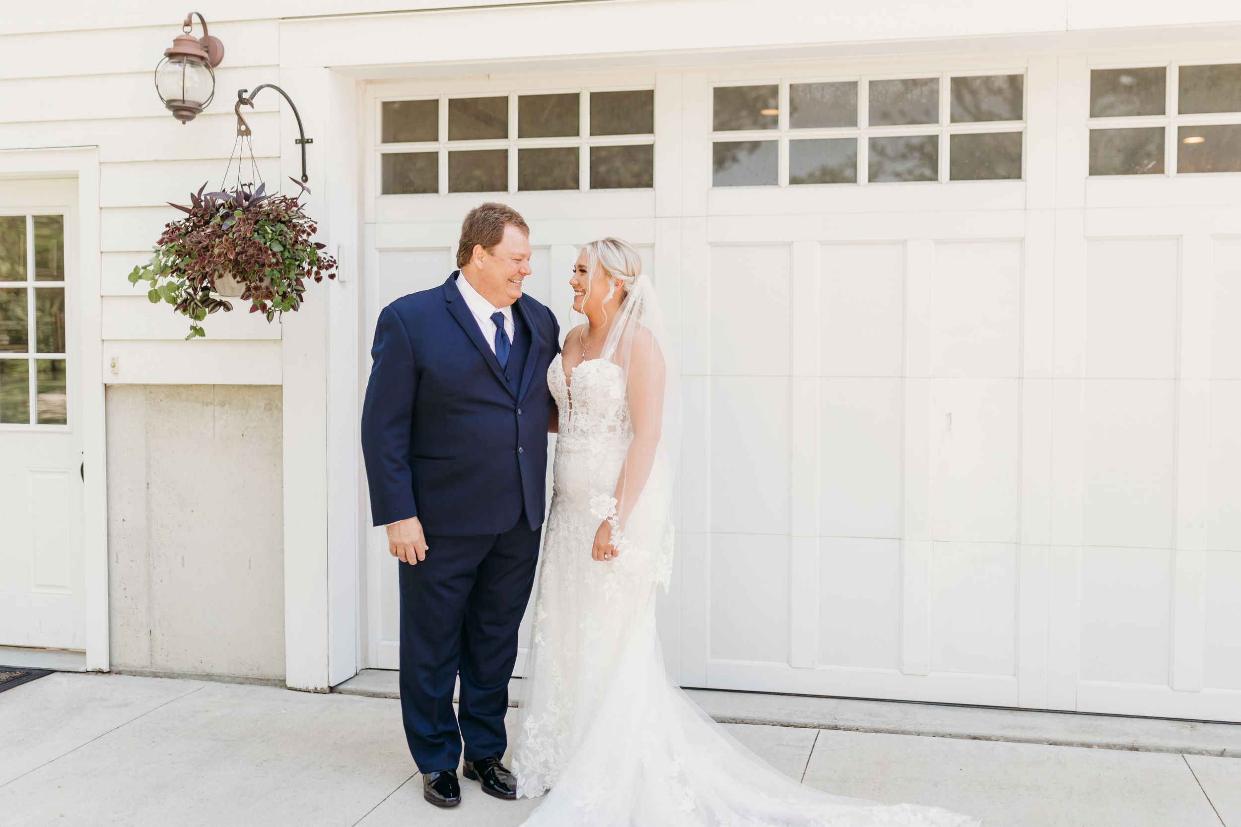 Bride in lace gown and groom in navy suit share a romantic moment outside a white barn wedding venue.