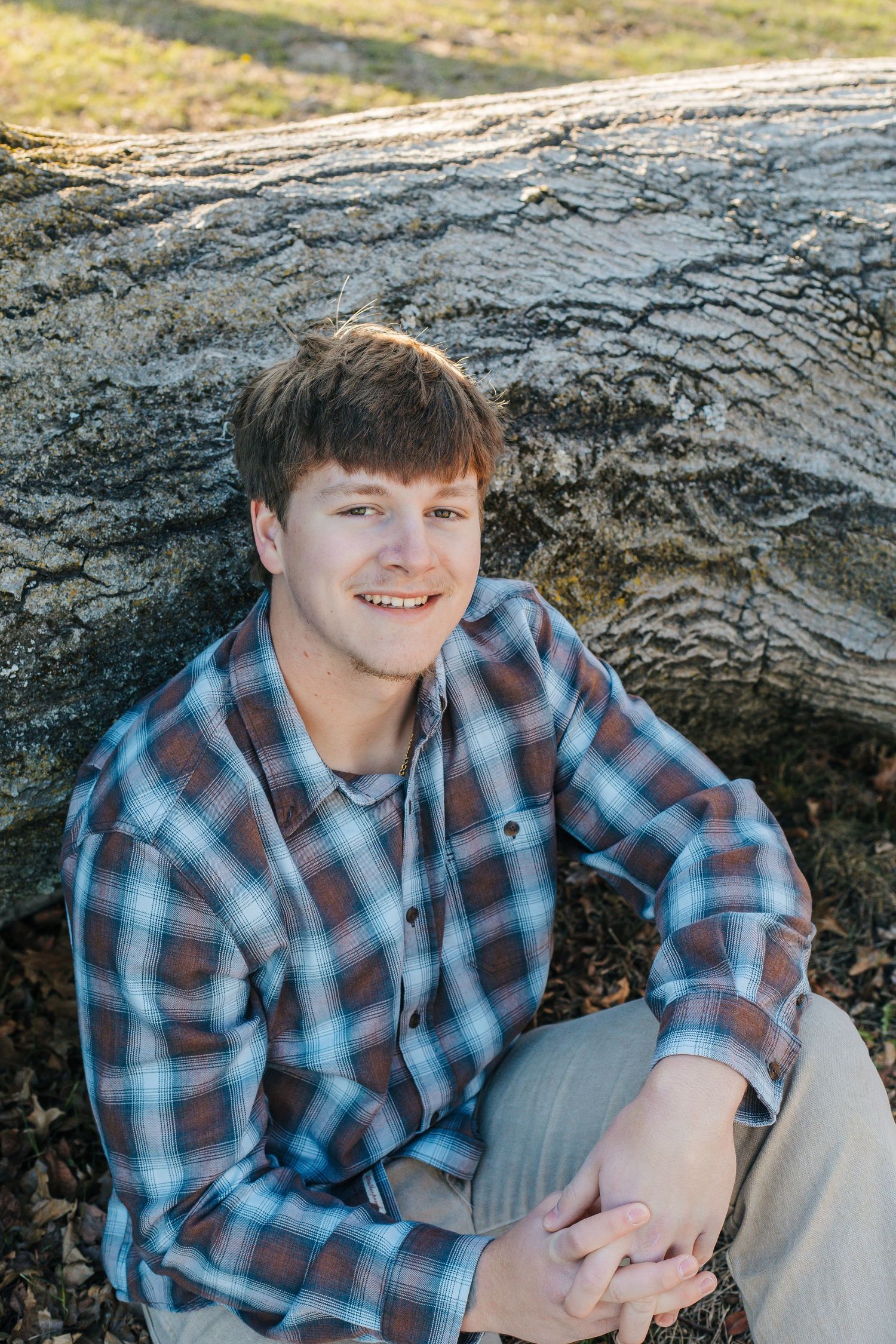 Smiling young man in plaid shirt sitting against a fallen log outdoors in natural light.