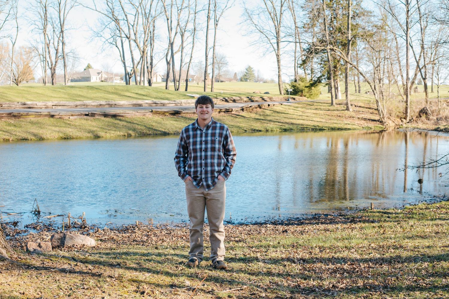 Young man in plaid shirt standing by a serene pond in a park on a clear day.