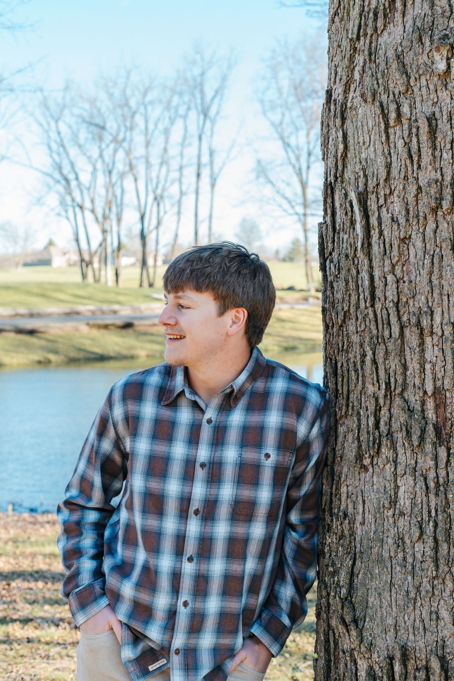 Young man in plaid shirt leaning against tree trunk near pond in park during autumn.