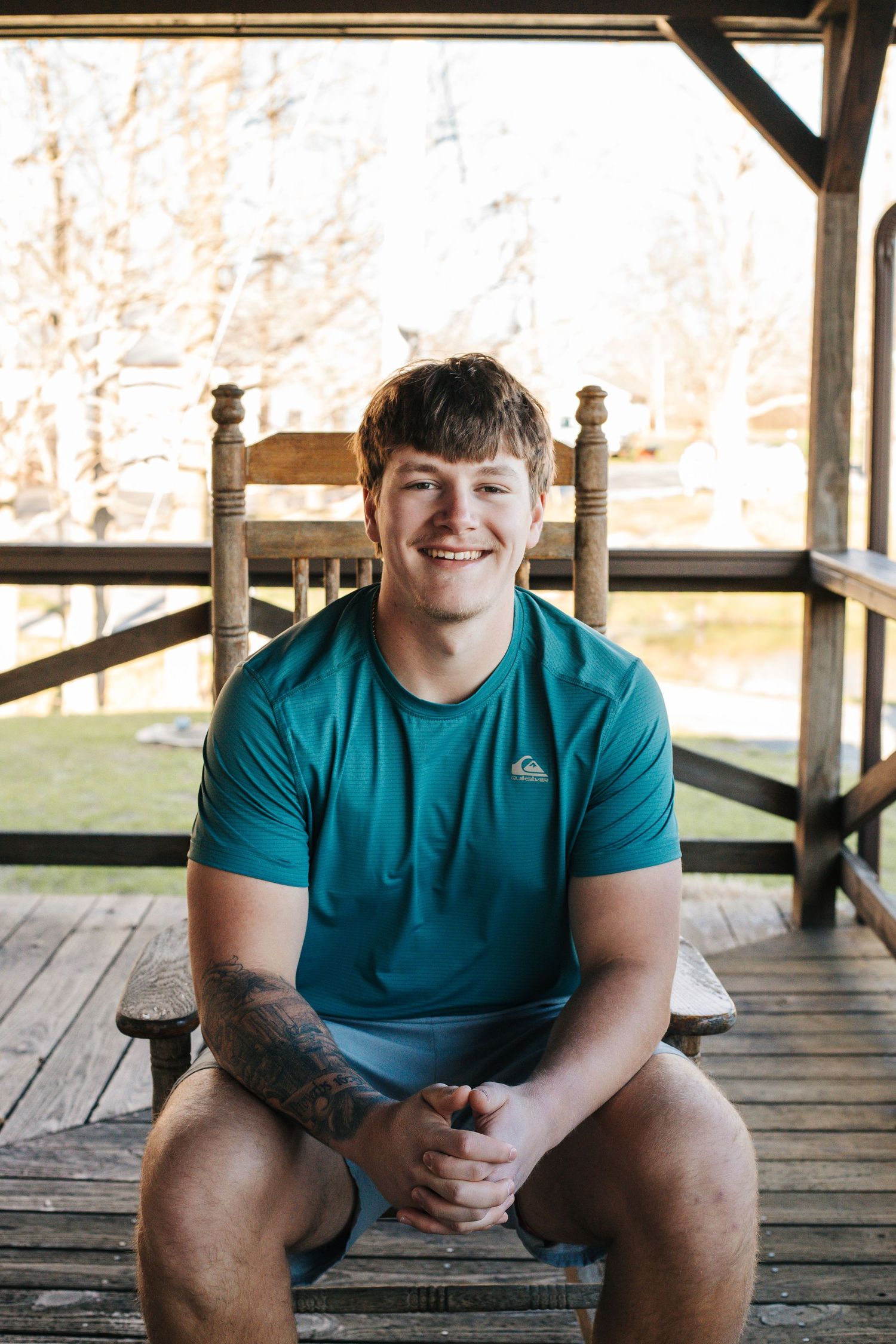 Smiling young man in teal Quiksilver shirt with tattoo sitting on wooden porch chair outdoors.