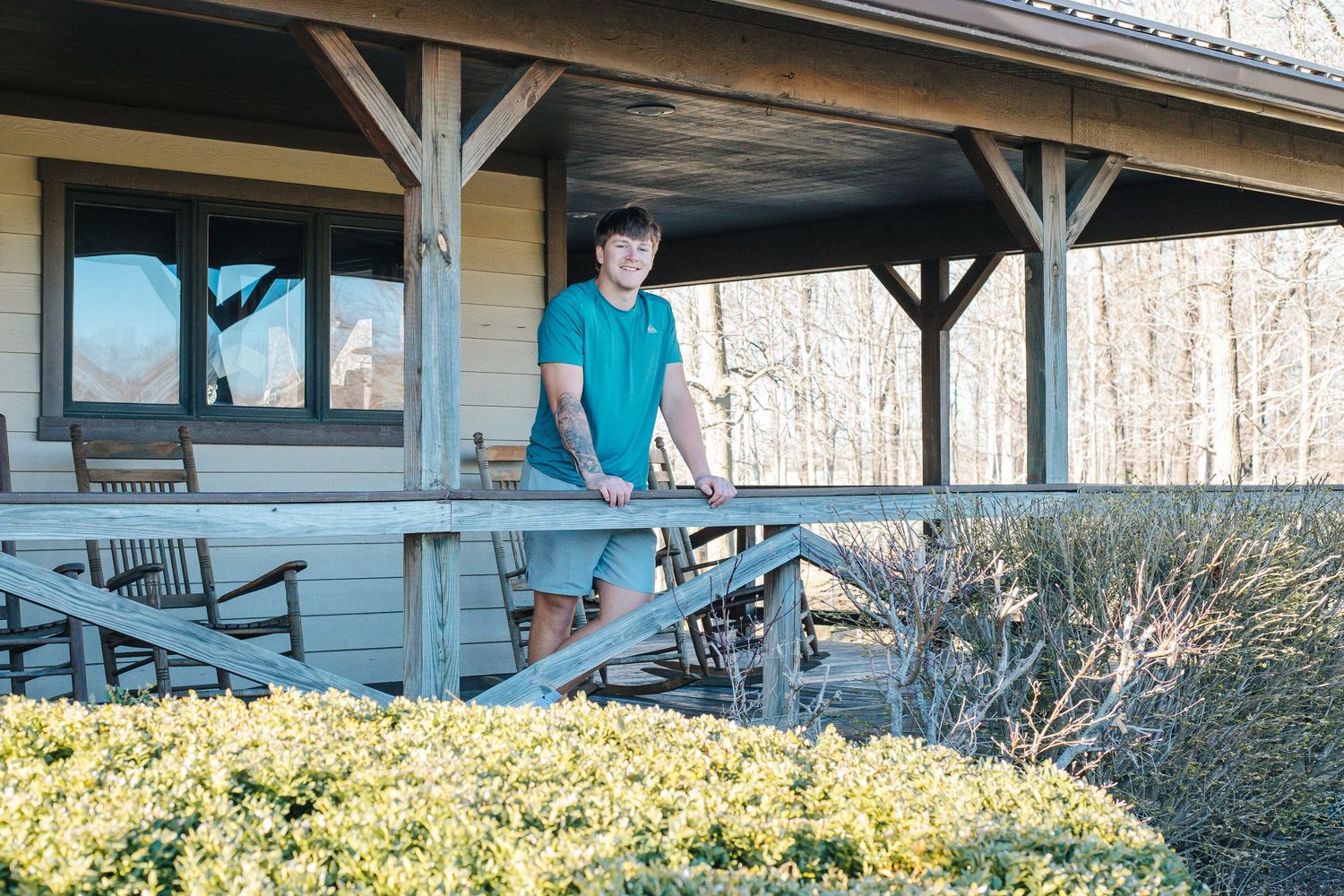 Senior Boy standing on wooden deck of rustic cabin surrounded by yellow shrubs in winter.