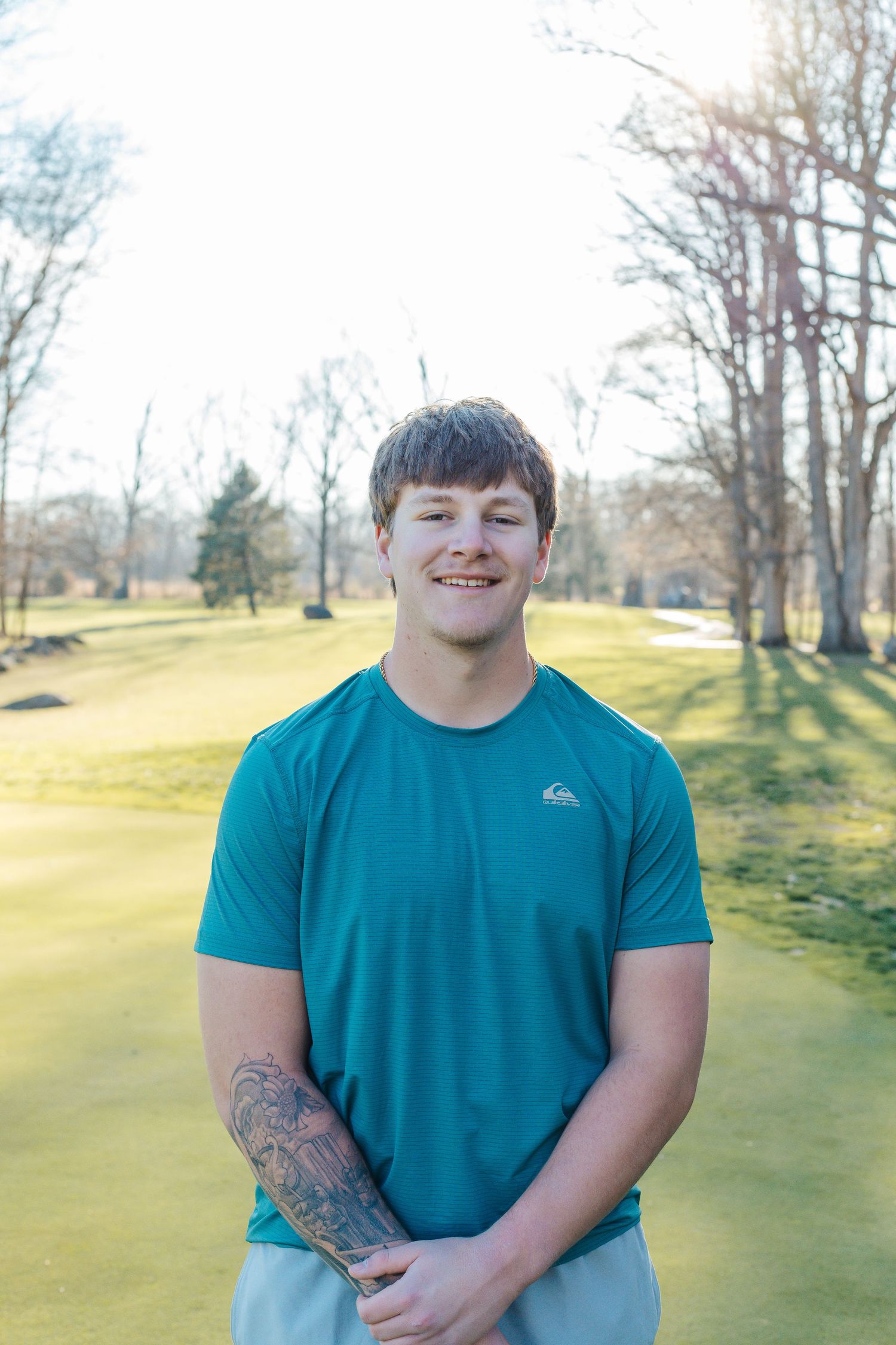 Smiling young man with tattoo sleeve wearing teal Quiksilver shirt standing on sunny golf course.
