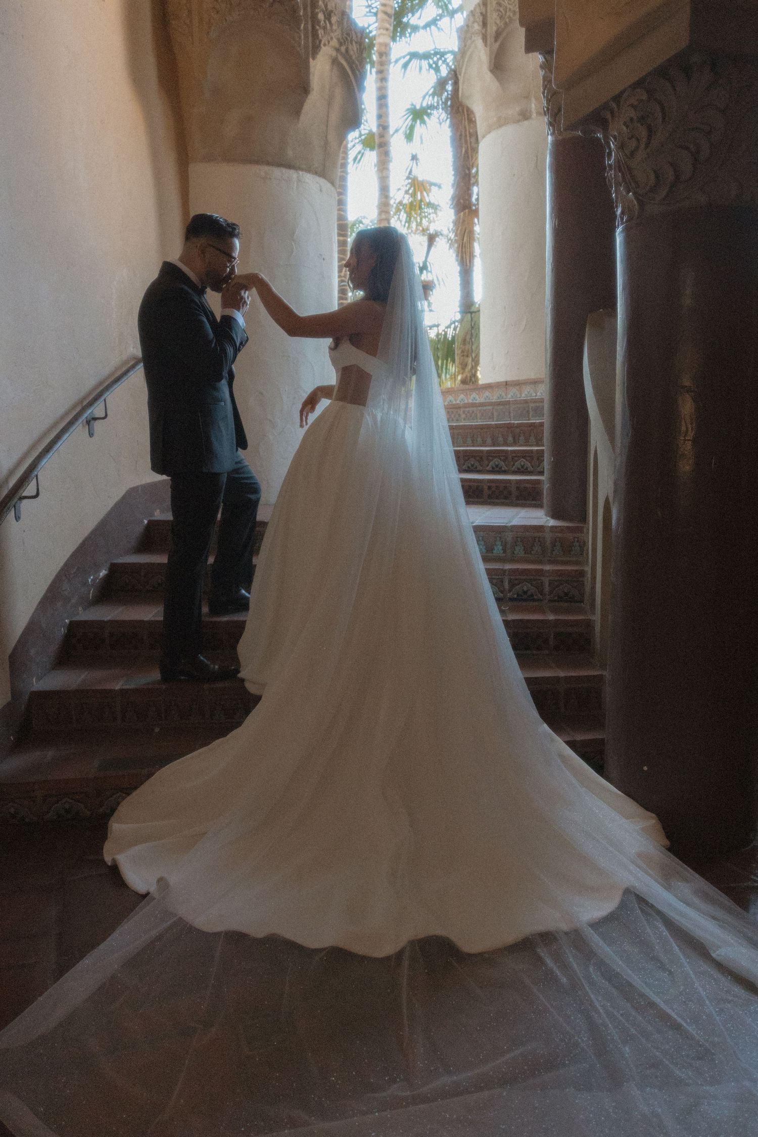Bride in flowing white gown and veil with groom in tuxedo sharing romantic moment on ornate staircase.
