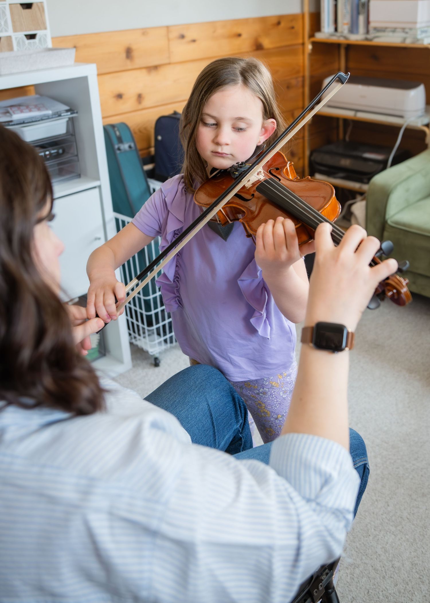 A young girl receives a violin lesson from a teacher in a cozy home music room setting.