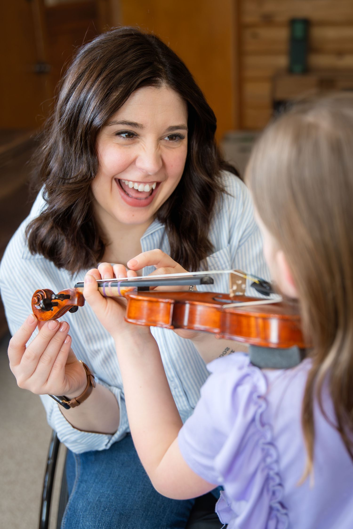 Woman in wheelchair smiling while teaching young girl to play violin indoors.